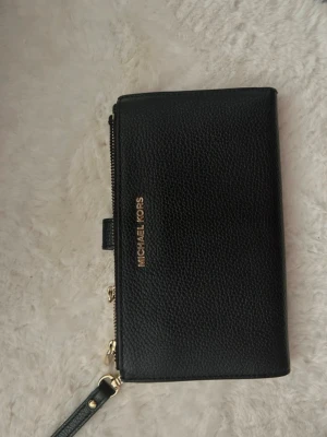Svart plånbok från Michael Kors - Stilren svart plånbok från Michael Kors i präglat läder med guldfärgade detaljer. Plånboken har flera kortfack, fönster för ID, dragkedjefack och avtagbar handledsrem. Perfekt för att hålla ordning på kort och kontanter.