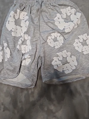 Grå shorts med vita blommor - Tqr emot motbud! Snygga grå shorts med elastisk midja och snörning. Shortsen har stora vita blommönster tryckta över hela plagget och är tillverkade i ett mjukt bomullsliknande material. Perfekta för en avslappnad och trendig stil.