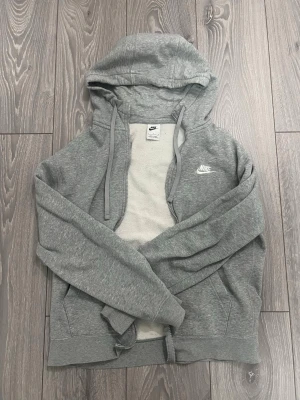 Grå zip hoodie från Nike - Snygg grå zip hoodie från Nike med vit logga på bröstet. Hoodien har dragsko i huvan, fickor framtill och mjukt fleecefoder på insidan. Perfekt för chill dagar eller när du vill ha en sportig look.