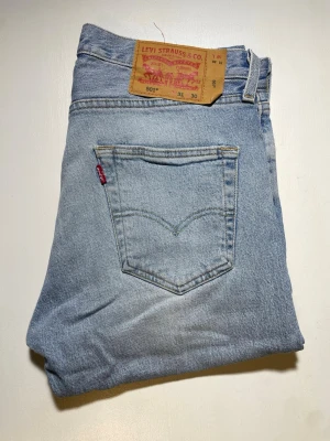 Levi's 501 ljusblå raka jeans - Klassiska Levi's 501 jeans i ljusblå tvätt med raka ben och femficksmodell. Jeansen har den ikoniska röda Levi's-lappen på bakfickan och patch i läder bak i midjan. Tillverkade i slitstark denim, perfekt för en avslappnad och tidlös stil.