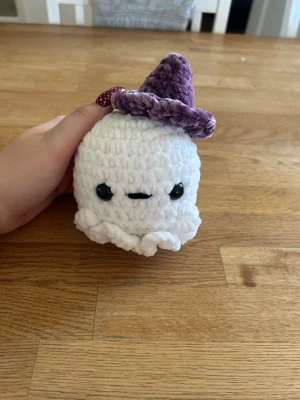 Virkad spöke med hatt - Söt handvirkad spöke i mjuk vit plysch med svarta säkerhetsögon. Spöket har en lila virkad hatt på huvudet och är perfekt som dekoration eller present. Liten och smidig att hålla i handen.