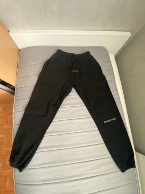 Svarta Essentials mjukisbyxor i storlek S - Svarta mjukisbyxor från Essentials med dragsko i midjan och resår vid bensluten. Byxorna har en enkel och clean look med Essentials-loggan tryckt på ena benet. Perfekta för chill dagar eller när du vill ha det bekvämt.