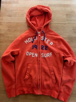 Hollister Hoodie  - Snygg orange hoodie från Hollister med dragkedja, blåa snören i huvan och broderad text 'Hollister 1922 Open Surf' i vitt och blått på bröstet. Två stora fickor framtill och mjukt material som känns skönt mot huden. Perfekt för en avslappnad stil. Skriv vid funderingar?