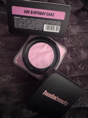 Huda Beauty Easy Bake Ube Birthday Cake - Lös setting powder i nyansen Ube Birthday Cake från Huda Beauty. Puder i en lila ton som ger en fräsch och jämn finish. Kommer i en fyrkantig burk med svart lock och rosa text. Perfekt för att fixera sminket och highlighta ansiktet.