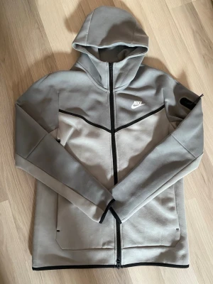 Grå Nike Tech Fleece hoodie - Snygg grå Nike Tech Fleece hoodie med dragkedja framtill och svart detaljlinje över bröstet. Har huva, två fickor fram och en extra ficka med dragkedja på ärmen. Tillverkad i mjukt och bekvämt material som passar perfekt till streetwear.