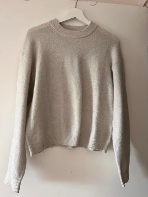 Beige stickad tröja med rund hals - Mysig beige stickad tröja med rund halsringning och ribbade muddar vid ärmslut och nederkant. Tröjan har en avslappnad passform och långa ärmar, perfekt för kyligare dagar.