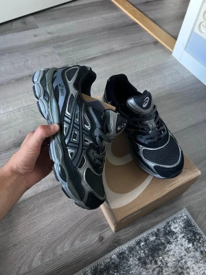 Svarta och grå Asics sneakers - Snygga sneakers från Asics i svart och grått med mesh- och syntetpaneler. Skorna har en chunky sula och klassisk snörning. Perfekta för dig som gillar sportig stil och vill ha bekväma skor till vardags.