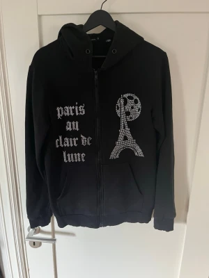 Svart hoodie med strasstryck Paris - Säljer en svart hoodie med dragkedja och huva. På framsidan finns ett strasstryck med texten 'paris au clair de lune' samt motiv av Eiffeltornet och månen i glittrande stenar. Hoodien har fickor framtill och ribbade muddar. Perfekt för dig som gillar statement-plagg och Paris-vibes.