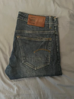 Blå jeans från G-Star RAW - Snygga blå jeans från G-Star RAW med klassisk femficksdesign och bruna detaljer på bakfickorna. Jeansen har en läderpatch med logga bak i midjan och är tillverkade i slitstarkt denim. Perfekta för en avslappnad och trendig stil.