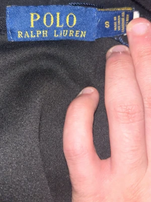 Svart hoodie från Polo Ralph Lauren - Säljer en svart hoodie från Polo Ralph Lauren med dragkedja framtill och klassisk vit logga på bröstet. Hoodien har justerbar huva med snören och en stor magficka. Perfekt för en chill och stilren look.