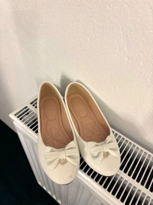 Vita ballerinaskor med rosett - Snygga vita ballerinaskor i konstläder med rund tå och dekorativ rosett framtill. Rosetten har en liten guldfärgad ring som detalj. Skorna har en mjuk innersula med blommönster för extra komfort. Perfekta för dig som gillar en söt och stilren look. Har en liten fläck de bild.