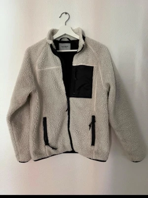 Vit teddyjacka från Carhartt - Säljer en vit teddyjacka från Carhartt med svarta detaljer. Jackan har dragkedja framtill, två sidofickor och en bröstficka i svart tyg. Materialet är mjukt och fluffigt, perfekt för kyliga dagar. Ståkrage och avslappnad passform.