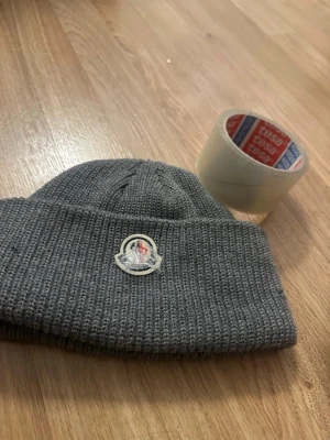 Grå ribbstickad mössa - Size L. Säljer en grå ribbstickad mössa från Moncler med uppvikt kant och klassisk Moncler-logga framtill. Mössan är i mjukt material och har en enkel, stilren design som passar till det mesta. Perfekt för kalla dagar och snygg till streetwear.
