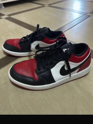 Svarta, röda och vita sneakers Jordan - Snygga sneakers i klassisk färgkombination av svart, rött och vitt. Modellen har lågt skaft, svart swoosh och perforerad tå. Skorna har svarta skosnören och vit sula med röd undersida. Perfekta för dig som gillar streetwear och sportig stil.