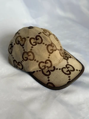 Äkta Gucci keps - Äkta Keps från Gucci med det klassiska GG-mönstret i en beige och brun färgskala. Kepsen har en justerbar rem baktill för passform. Storlek S 56cm