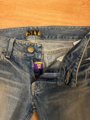 Blå slitna jeans från SLY - Ett par blå jeans från SLY med snyggt sliten tvätt och coola detaljer som lila etiketter med texten 'COME ON IN' och 'WELCOME'. Klassisk femficksmodell med dragkedja och knapp. Perfekta för en avslappnad streetstil.