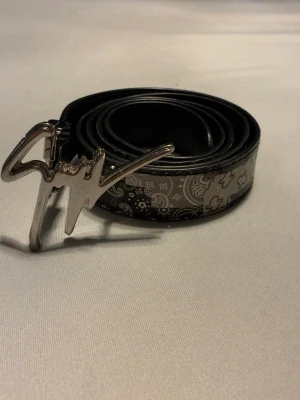 Svart bälte från giuseppe  - Snyggt svart bälte med paisleyinspirerat mönster och en unik silvrig buckle i form av en signatur. Perfekt för att lyfta vilken outfit som helst och ger en cool touch till jeans eller byxor.