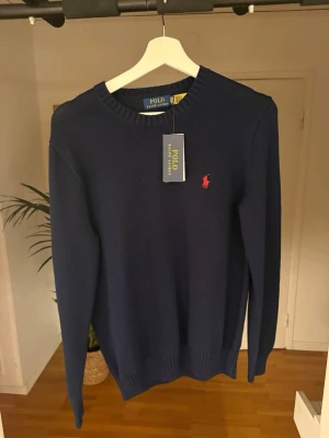 Mörkblå stickad tröja Polo Ralph Lauren - Mörkblå stickad tröja från Polo Ralph Lauren med rund halsringning och röd broderad logga på bröstet. Tröjan har ribbstickade muddar vid ärmslut och nederkant. Perfekt för en stilren och klassisk look.