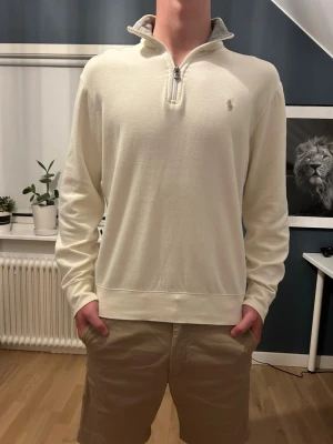 Vit half-zip tröja från Polo Ralph Lauren - Snygg vit half-zip tröja från Polo Ralph Lauren med grå insida på kragen och broderad logga på bröstet. Tröjan har en hög krage med dragkedja och är tillverkad i mjukt material som känns skönt mot huden. Perfekt för en clean och stilren look.
