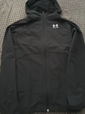Svart vindjacka från Under Armour - Snygg svart vindjacka från Under Armour med huva och hel dragkedja framtill. Jackan har en minimalistisk design med logga på bröstet och är tillverkad i ett lätt syntetmaterial som passar perfekt för blåsiga dagar.