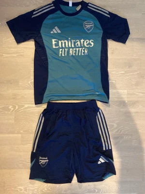 Arsenal träningsset blå/turkos Adidas - Säljer ett Arsenal träningsset från Adidas i blått och turkost. Setet består av en t-shirt och shorts med klubbmärke och Adidas-logga. Snygga vita detaljer och tryck på både tröja och shorts. 💨☺️