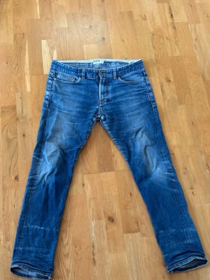 Blå raka jeans med slitningar - Säljer ett par klassiska blå jeans med raka ben och snygga slitningar framtill och baktill. Jeansen har fem fickor, normal midja och är tillverkade i ett mjukt denimtyg. Perfekta för en avslappnad och trendig look.