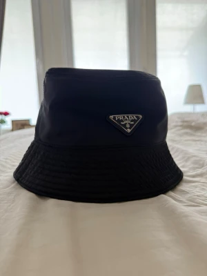 Svart bucket hat från Prada - Snygg svart bucket hat från Prada med klassisk trekantig logotyp framtill. Hatten är tillverkad i ett glansigt material som ger en lyxig känsla och har en stilren, enkel design som passar till många outfits.