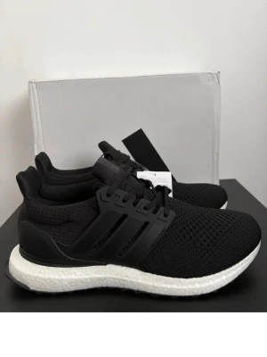 Svarta Adidas Ultraboost sneakers - Svarta Adidas Ultraboost sneakers med stickad ovandel i mesh och klassiska tre ränder på sidan. Skorna har en vit Boost-sula som ger grym komfort och respons. Perfekta för dig som gillar sportig stil och vill ha något som funkar till det mesta.