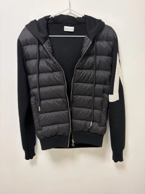 Moncler jacka  - Snygg svart pufferjacka från Moncler med stickade ärmar och stor vit M-detalj på ena ärmen. Jackan har huva, dragkedja framtill och ribbade muddar.