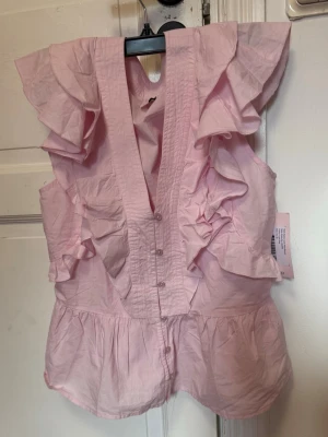 Rosa blus med knappar - Supersöt rosa blus med V-ringning och volanger längs axlar och framstycke. Blusen är ärmlös och har knäppning framtill. Den är inte alls så genomskinlig som den ser ut. Ovanpå ser den ut som på första bilden, så det är inget att oroa sig över. Jag säljer den eftersom den inte passar och priset går att diskutera. Helt sprillans ny och aldrig använd med prislappen kvar. 💕
