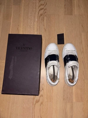 Valentino open Garavani vita sneakers - Snygga vita sneakers från Valentino Garavani med bred marinblå rem över vristen och klassisk snörning. Skorna har en stilren design med diskret logga på plösen och detaljer i vitt och marinblått. Tillverkade i skinn för en exklusiv känsla. Dem är använda som ni ser därav priset. Priset är ej hugget i sten, om ni har frågar så är de bara att fråga🤗