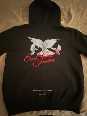 Svart hoodie från Hoodrich med tryck - Svart hoodie med huva från Hoodrich. Stora tryck med vita duvor och texten 'From Nothing To Something' på ryggen samt 'HOODRICH' och duvor på framsidan. Känguruficka framtill och detaljer i rött och vitt. Perfekt streetwear-stil.