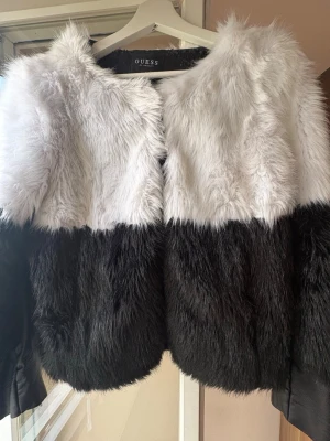 Svartvit pälsjacka från Guess - Säljer en fluffig pälsjacka från Guess i svart och vitt. Jackan har en rak passform, rund halsringning och är tillverkad i mjukt fuskpälsmaterial. Färgen är blockad med vit upptill och svart nedtill. Perfekt för dig som vill sticka ut i vinter!