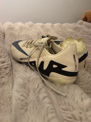 Nike Mercurial Air Zoom fotbollsskor - Säljer ett par gräddvita Nike Mercurial Air Zoom fotbollsskor med svart swoosh och detaljer. Skorna har snörning, låg profil och dobbar för bra grepp på planen. Tillverkade i syntetmaterial för lätt känsla och snabbhet.
