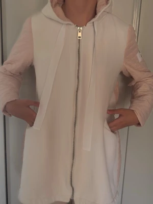 Ljusrosa lång hoodiejacka med dragkedja - Lång hoodiejacka i ljusrosa och vitt med dragkedja framtill, stora fickor och justerbart band i midjan. Jackan har quiltade ärmar och baksida samt en stor huva. Perfekt för lager på lager och chill stil.