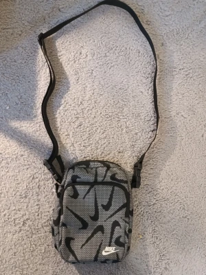 Nike shoulder bag 160kr - Nike shoulder bag 160kr Titel: Nike Heritage axelremsväska / crossbody (Swoosh-mönstrad) Beskrivning: Säljer en praktisk och snygg liten axelremsväska (crossbody) från Nike med heltäckande Swoosh-mönster. Perfekt storlek för mobil, plånbok, nycklar och andra småsaker. Märke: Nike Modell: Crossbody / Axelremsväska Färg/Mönster: Grå/svart med heltäckande Nike-mönster och vit broderad logga Fack: Stort huvudfack samt ett praktiskt ytterfack med dragkedja Detaljer: Justerbar axelrem för perfekt pass