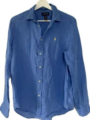 Blå linneskjorta från Polo Ralph Lauren - Klassisk blå skjorta i linne från Polo Ralph Lauren med ikonisk gul broderad logga på bröstet. Skjortan har lång ärm, knappar framtill och en avslappnad passform. Perfekt till jeans eller chinos för en clean look.