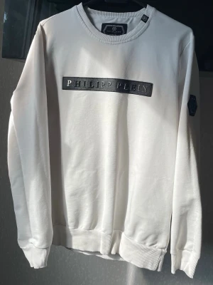 Vit sweatshirt från Philipp Plein - Säljer en stilren vit sweatshirt från Philipp Plein med svart logotyp framtill och en cool patch på ärmen. Tröjan har rund halsringning, ribbade muddar och är tillverkad i mjukt material. Perfekt för dig som gillar exklusiva streetwear-plagg.