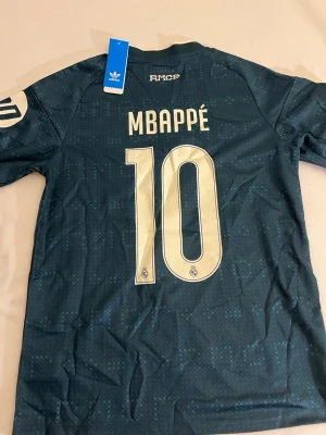 Real Madrid Mbappé matchtröja - Snygg mörkblå Real Madrid matchtröja från Adidas med Mbappé och nummer 10 på ryggen. Tröjan har vita detaljer, klubbmärke och sponsortryck framtill samt Champions League-märke på ärmen. Tillverkad i lätt och ventilerande material.