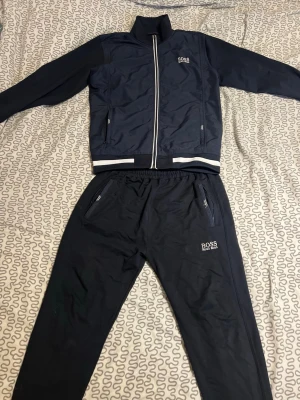 Mörkblått set från BOSS - Snyggt mörkblått set från BOSS med vit dragkedja och vita detaljer vid ärmslut och midja. Setet består av en långärmad zip-jacka och matchande byxor med resår i midjan och dragkedjefickor. BOSS-logga på både jacka och byxa. Perfekt för en sportig och avslappnad look.