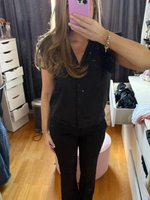 Svart blus med volangärmar - Snygg svart blus med knappar framtill och v-ringning. Blusen har volangprydda ärmlösa axlar i broderad spets. Aldrig använd.