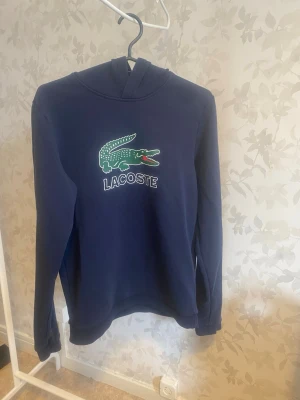 Mörkblå hoodie från Lacoste - Snygg mörkblå hoodie från Lacoste med det klassiska krokodilloggan broderad på bröstet. Hoodien har huva och långa ärmar. Perfekt för dig som gillar stilrena och sportiga plagg. Materialet känns mjukt och bekvämt.