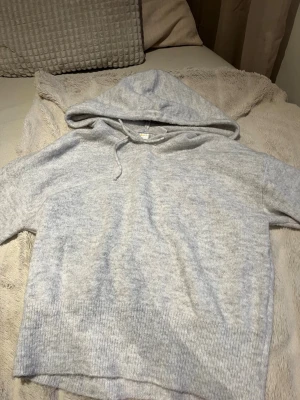 Ljusgrå hoodie från Gina Tricot - Mysig ljusgrå hoodie från Gina Tricot i mjukt stickat material. Tröjan har en stor huva, dragsko och bred ribbad kant nedtill. Perfekt för chill dagar och enkel att matcha med jeans eller leggings.