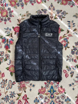 Svart dunväst från EA7 Emporio Armani - Snygg svart dunväst från EA7 Emporio Armani med quiltad design och hög krage. Västen har dragkedja framtill och logga på bröstet. Perfekt för lager-på-lager och ger en sportig vibe till din outfit.