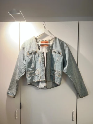 Ljusblå cropped jeansjacka Brooker - Trendig ljusblå jeansjacka från Brooker med croppad passform och rå kant nertill. Jackan har dekorativa dragkedjor framtill, långa ärmar och en justerbar snörning i ryggen. Perfekt för dig som vill ha en unik och cool denimlook.