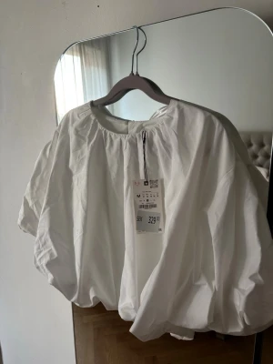 Vit puffig blus med kort ärm - Säljer en vit blus med puffiga, korta ärmar och rund halsringning. Blusen har en luftig och oversized passform med ballongliknande siluett. Tillverkad i ett lätt tyg som ger en fräsch och trendig look. Perfekt att styla med jeans eller kjol.
