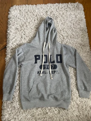 Grå hoodie med POLO-tryck -  Säljer en Ralph Lauren hoodie i storlek M men är för liten för mig så jag skulle säga att den är i storlek S och passar folk som är upp till 175 cm långa. Priset är ej hugget i sten