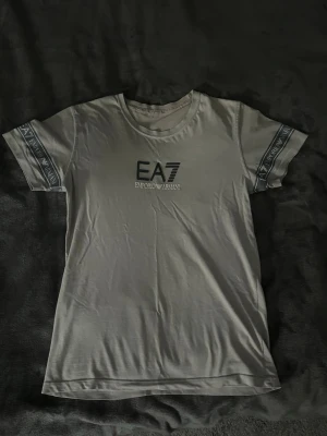 Vit EA7 Emporio Armani t-shirt - Snygg vit t-shirt från EA7 Emporio Armani med tryck på bröstet och logoband på ärmarna. Klassisk rund halsringning och korta ärmar. Perfekt för dig som gillar stilrena och trendiga plagg med exklusiv känsla.