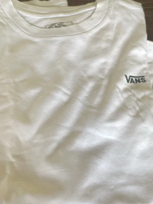 Vit t-shirt från Vans - Klassisk vit t-shirt från Vans med liten logga tryckt på bröstet. T-shirten har rund halsringning och är tillverkad i mjuk bomull som känns skön mot huden. Perfekt basplagg för alla tillfällen och enkel att matcha med andra plagg. Storlek L i barn