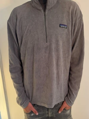 Grå fleece tröja från Patagonia - Mjuk och skön grå fleece tröja från Patagonia med halv dragkedja framtill och hög krage. Perfekt för kyliga dagar och enkel att slänga på sig. Diskret logga på bröstet och avslappnad passform.